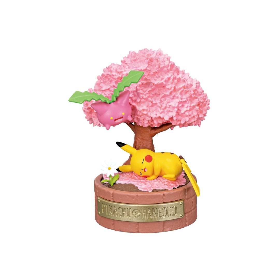 A small Pokémon figure featuring Pikachu sleeping under a pink cherry blossom tree while Hoppip floats above. The scene includes scattered petals, a white flower, and a round brick-patterned base labeled “PIKACHU & HANECCO.”